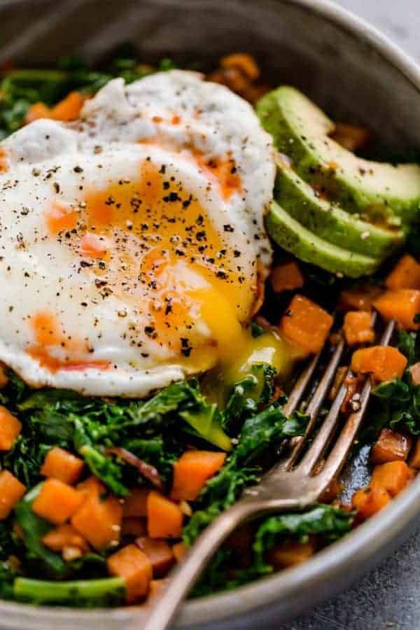 Close up photo taken at an angle of the Kale and Sweet Potato Sauté in a bowl with a fried egg on top with yolk running out of it and avocado slices.