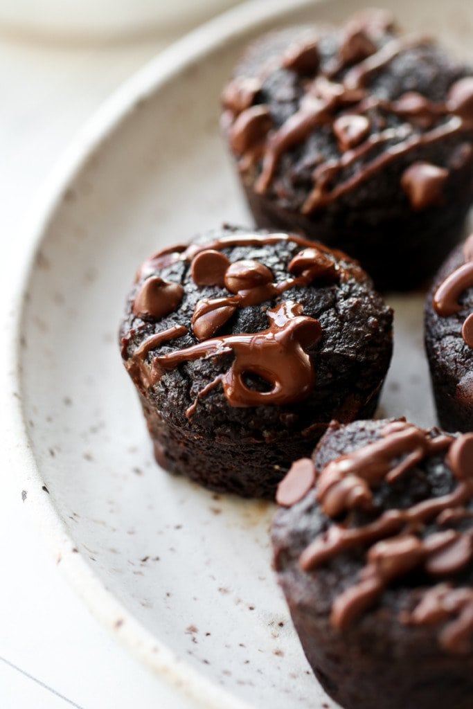 Close up view of a grain-free sweet potato avocado brownie bite topped with melted mini chocolate chips and chocolate drizzle, on a speckled plate with more brownie bites.