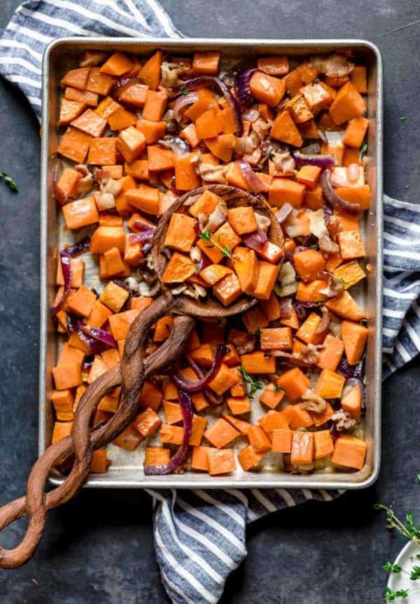 A large wooden spoonful of Roasted Sweet Potatoes with Maple and Bacon resting on a baking sheet full 