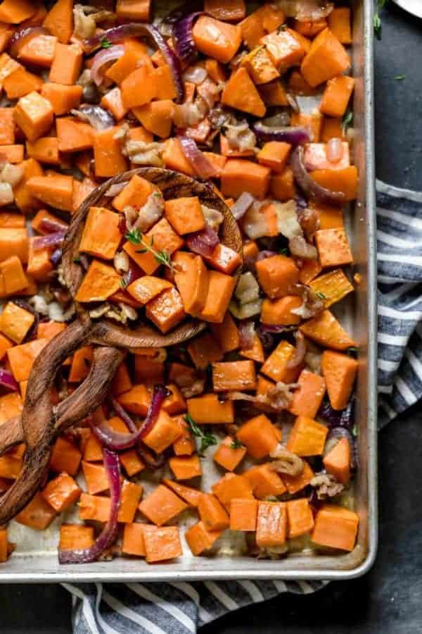 overhead view of a wooden spoonful of Roasted Sweet Potatoes with Maple and Bacon resting on a sheet pan of them