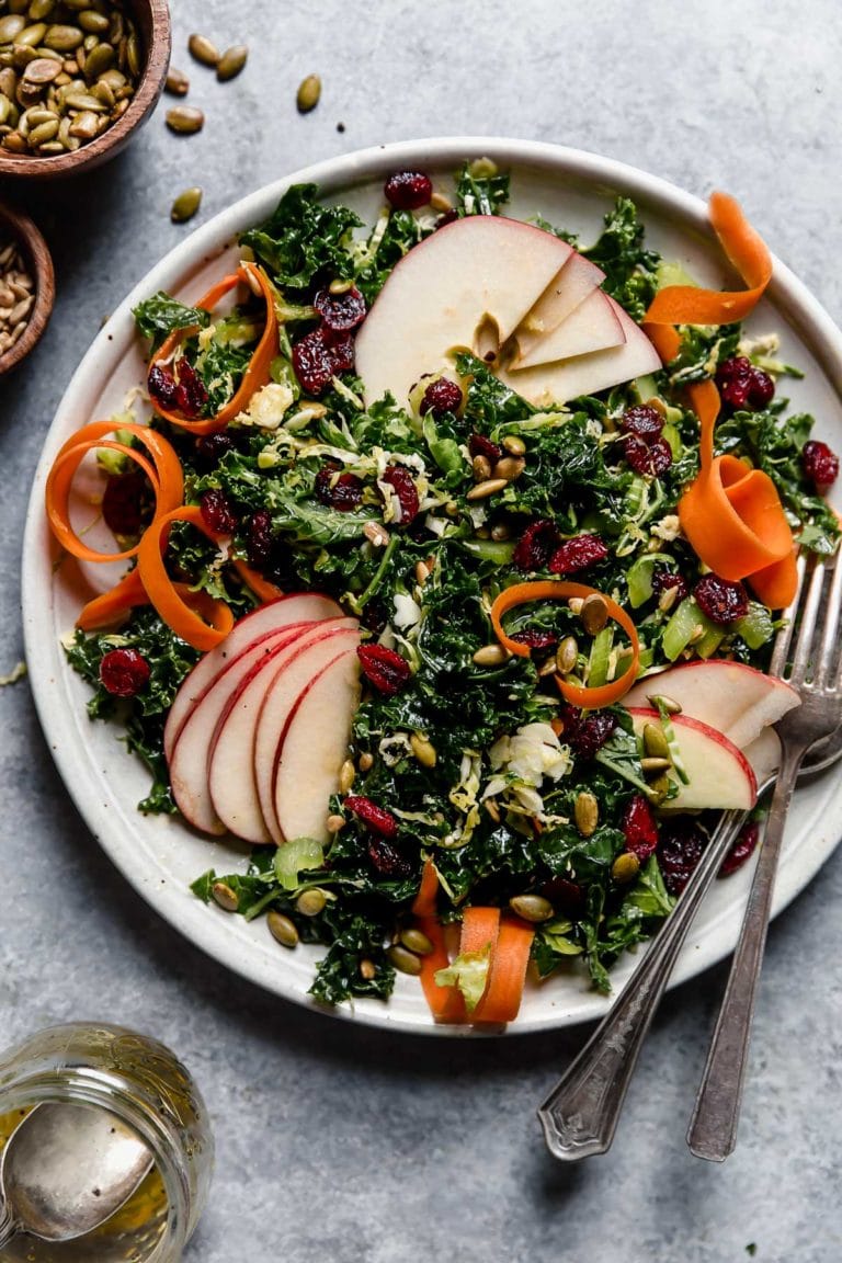 Overhead view of a plate of massaged kale salad topped with diced apples, celery, carrots, shredded brussels sprouts, dried cranberries, and pumpkin seeds. 