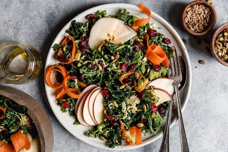 Kale salad on a white plate with dried cranberries, pepitas, apple slices, and carrot ribbons.