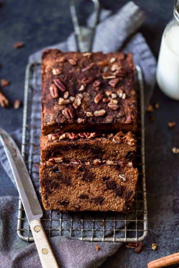 A loaf of gluten-free pumpkin bread with chocolate chips topped with pecans on a wire cooling rack being cut into thick slices.