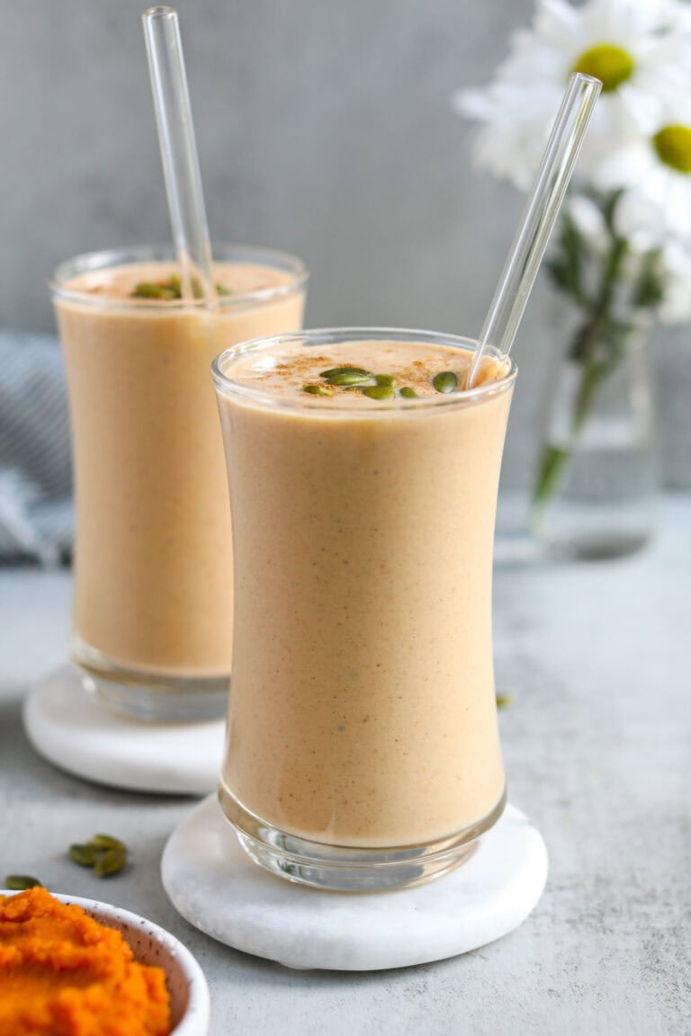 Two glasses full of protein pumpkin smoothie topped with pepitas, glass straws sticking out.