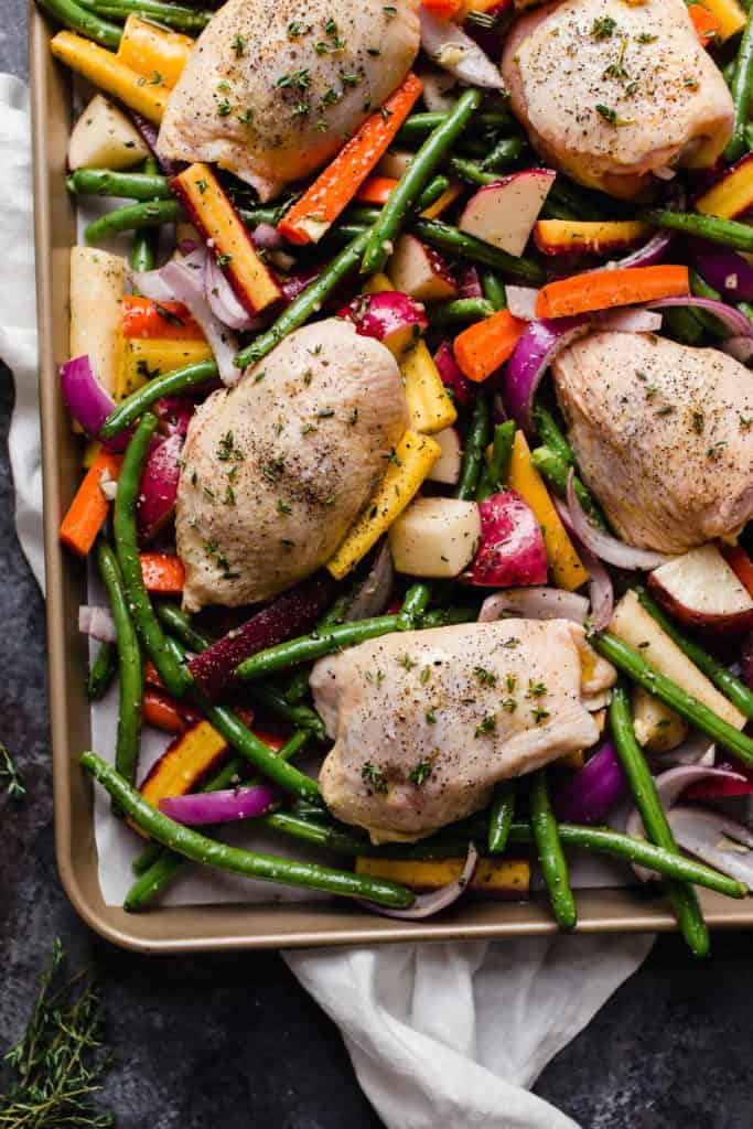 Overhead view raw chicken thighs on colorful veggies and quartered potatoes on baking sheet ready for baking.