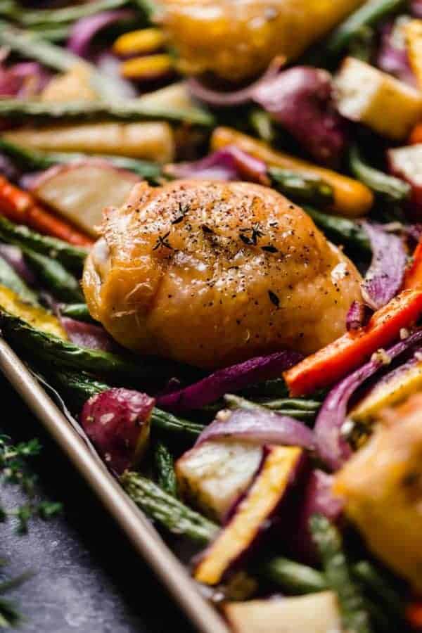 Close up side view baked chicken thigh on roasted vegetables and potatoes on baking sheet.