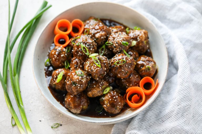 White bowl filled with meatballs coated in teriyaki sauce and garnished with sliced carrots and topped with sesame seeds. 