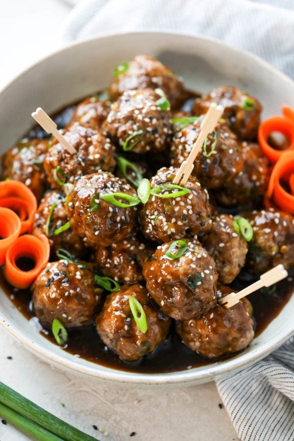 Teriyaki meatballs piled high in a bowl topped with sesame seeds, scallions, and serving toothpicks.