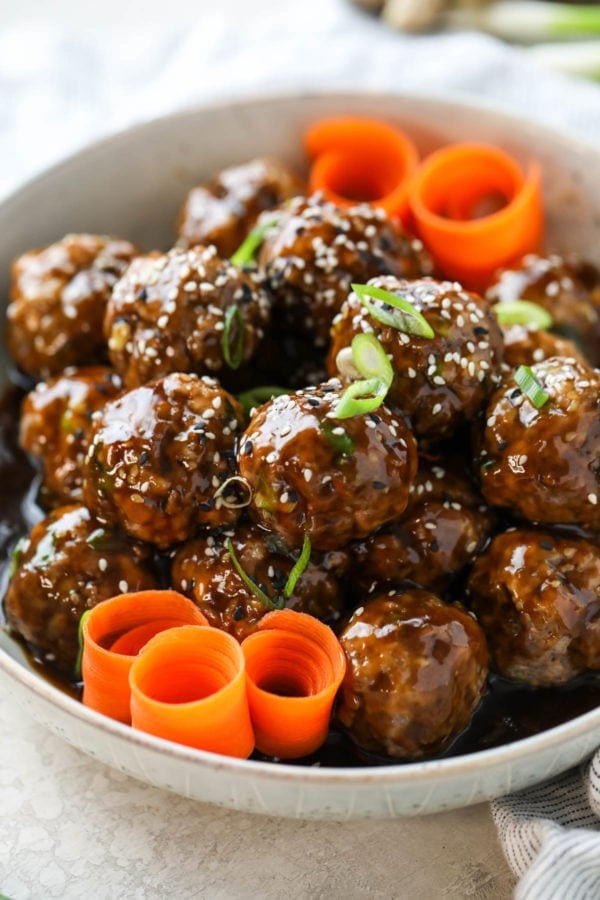 Teriyaki meatballs served in a white bowl topped with sesame seeds, scallions, and carrots swirls. 