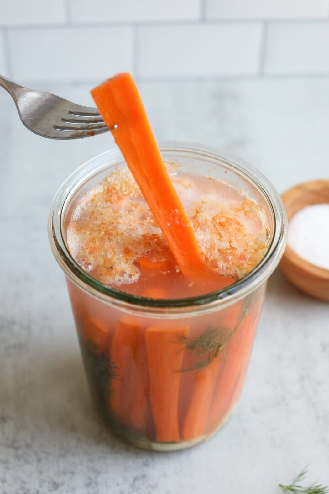 A fork lifting a fermented carrot stick from a jar. 