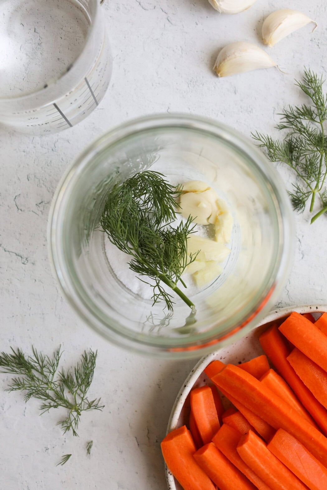 Garlic pieces and fresh herbs in a container, a plate of carrot sticks next to it. 