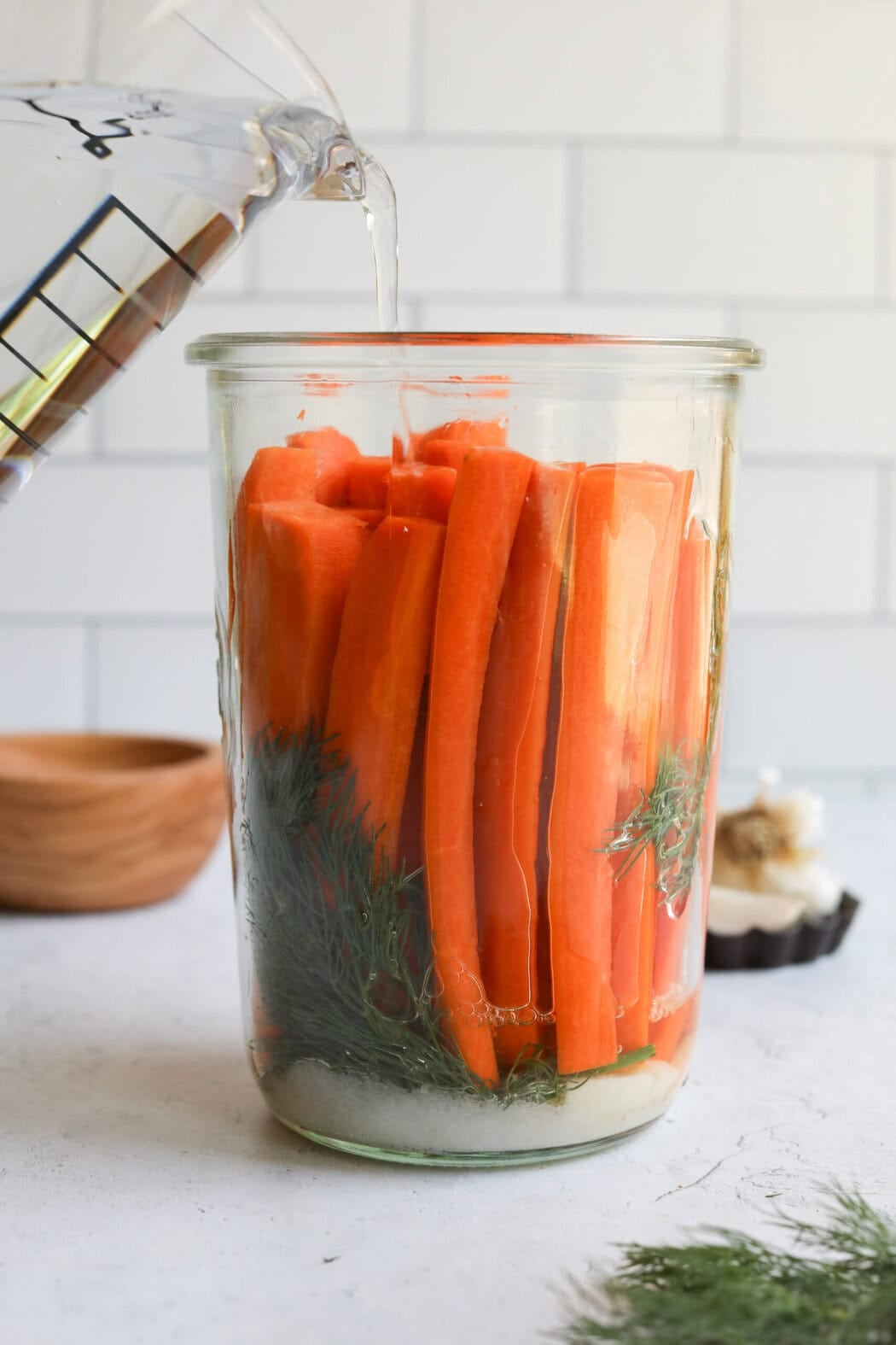Pickling liquid being poured into a jar  of carrot sticks and herbs. 