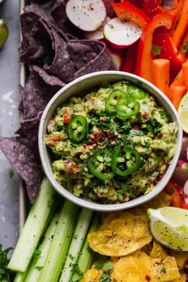 Overhead photo of Homemade Guacamole in a white bowl with a variety of colorful vegetables around the bowl as well as blue corn chips and plantain chips.
