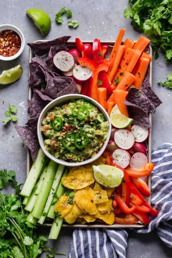 Overhead photo of Homemade Guacamole in a white bowl with a variety of colorful vegetables around the bowl as well as blue corn chips and plantain chips.
