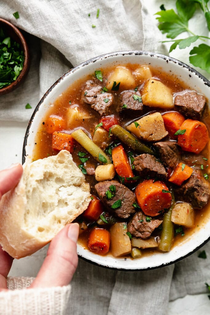 A rustic bowl filled with slow cooker beef stew with carrots, parsnips, and potatoes. A piece of crusty bread being dipped into soup.