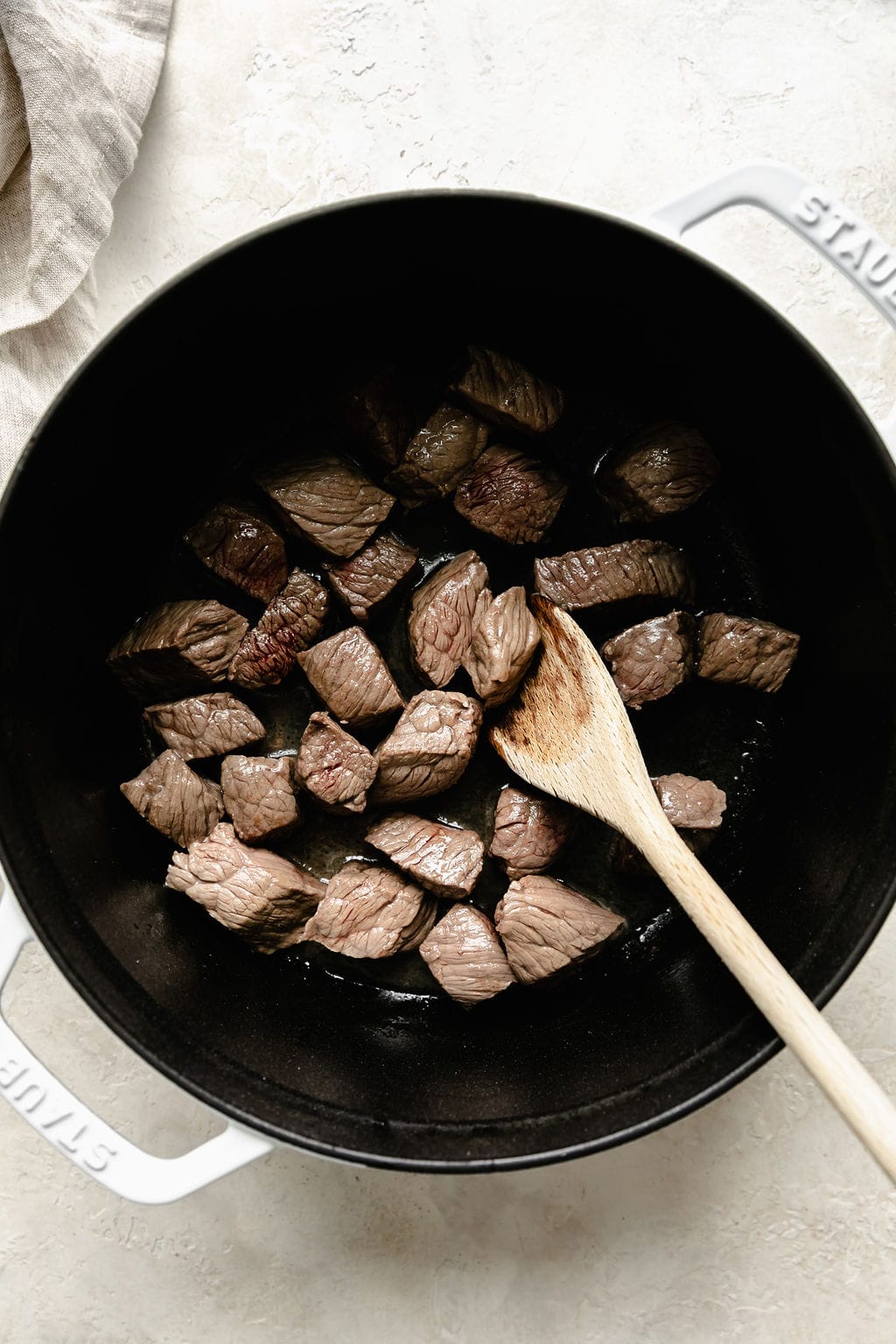 A wooden spoon stirring beef pieces in Staub skillet