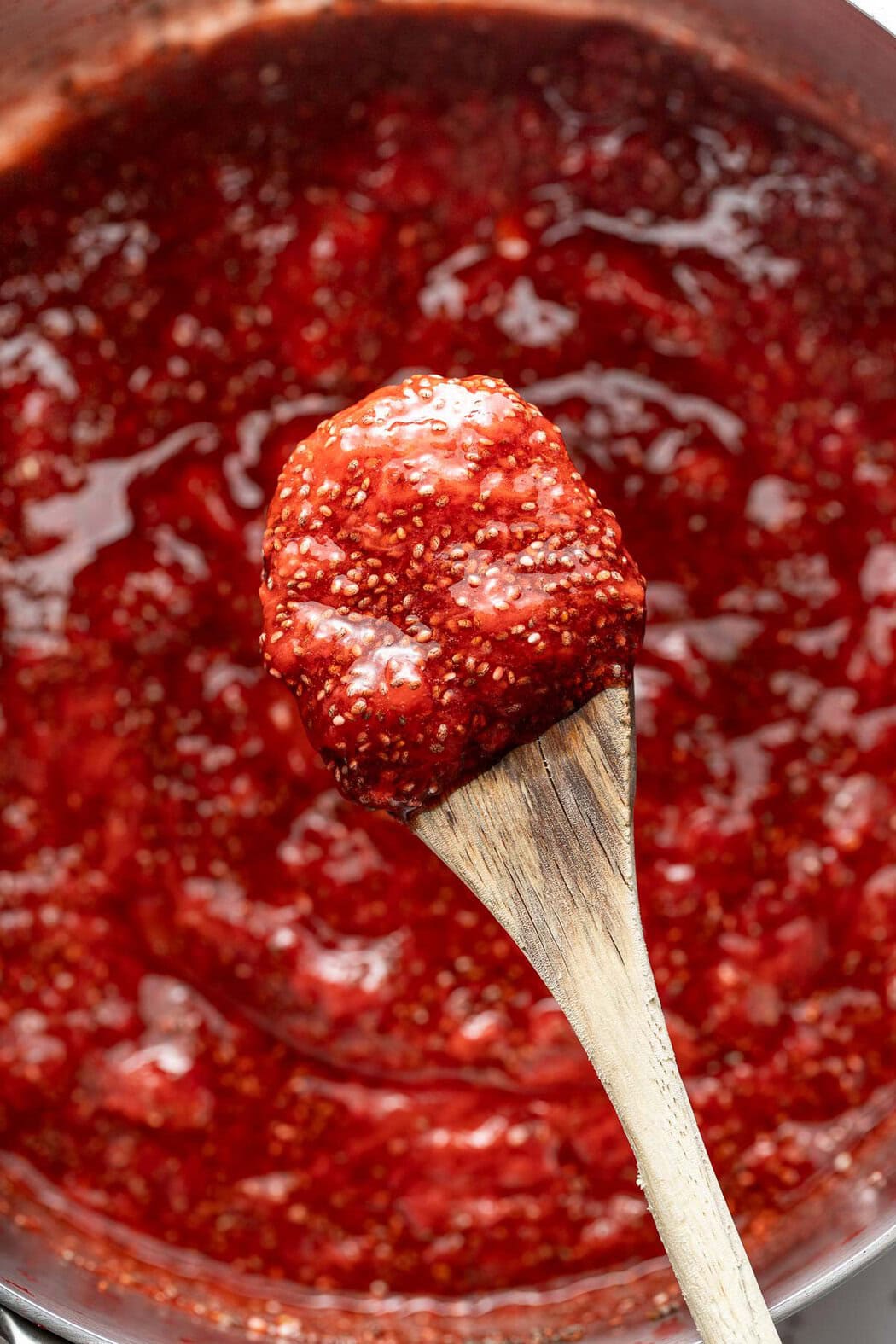 Overhead view of a spoonful of Chia Seed Jam in a wooden spoon over a pot filled with the strawberry mixture.