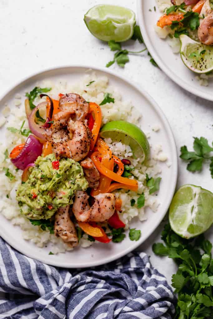 Chicken Fajitas on plate with cauliflower rice, peppers, onions, lime and guacamole on a white surface