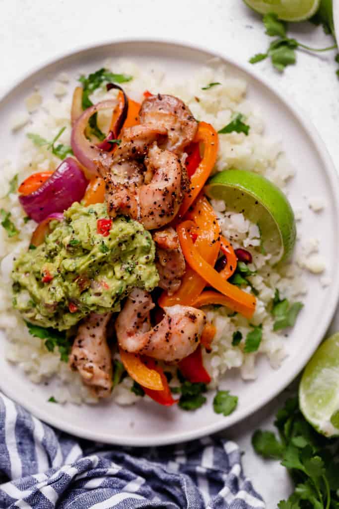 Close-up of chicken rajitas on plate with cauliflower rice, peppers, onions, lime and guacamole on a white surface