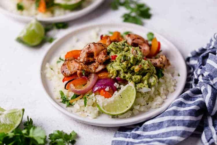 Chicken fajitas served over cauliflower rice on white plate with guacamole and lime wedges.