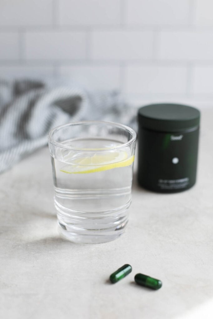 Glass of water with a slice of lemon floating inside it, seed brand probiotic pills next to the base of the glass. 