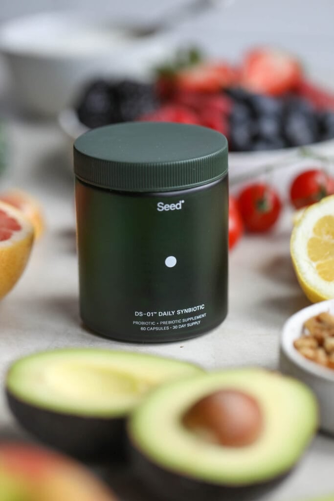 A canister of Seed probiotics with fresh fruits, nuts, and vegetables around it.