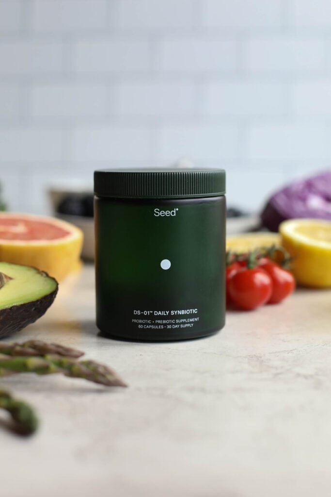 Canister of Seed probiotics on counter with fresh fruits and vegetables around it