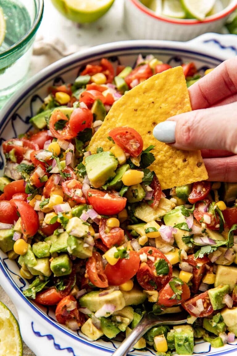 Close up view of a hand holding a chip dipping into a bowl of fresh avocado salsa topped with fresh cilantro.