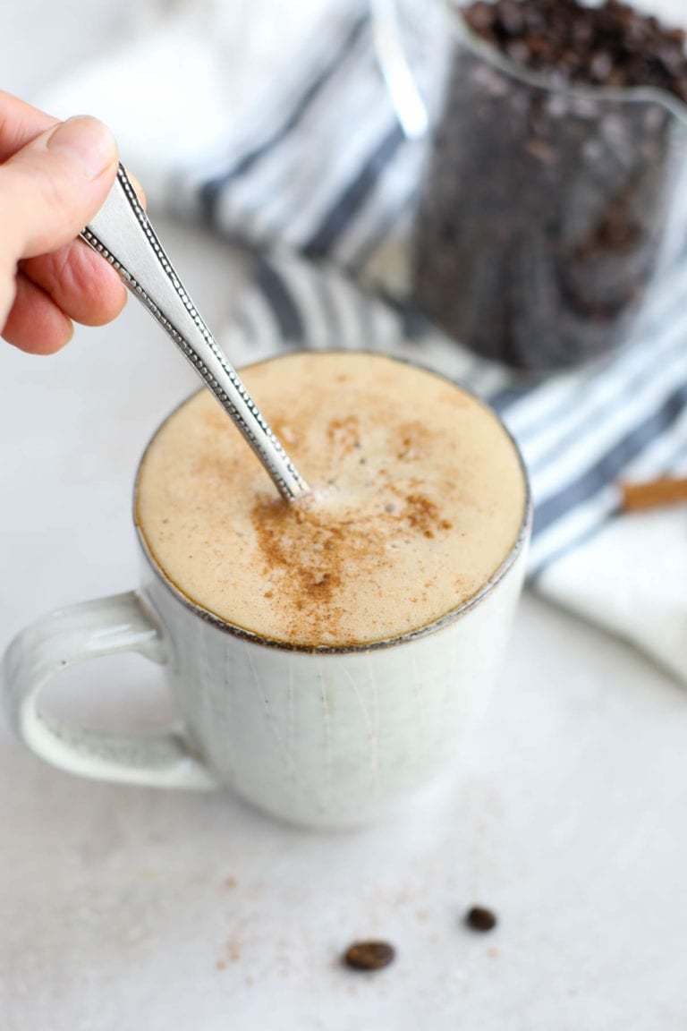 Cinnamon Coconut Latte in a cream mug with froth on top and a spoon stirring the liquid