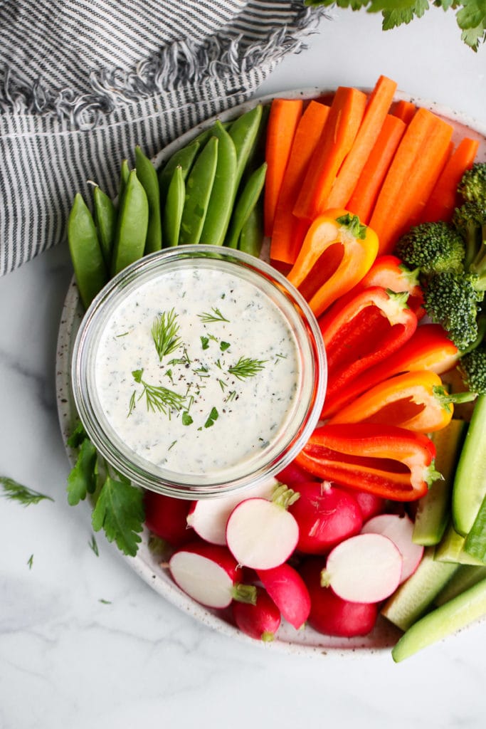 A platter filled with brightly colored veggies displayed around a small bowl of Paleo ranch dressing, topped with fresh dill