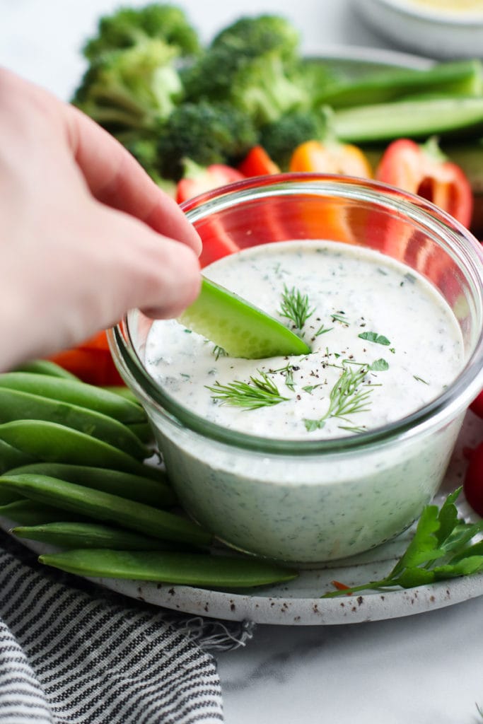 A fresh cucumber spear being dipped into a bowl of creamy homemade ranch dressing.