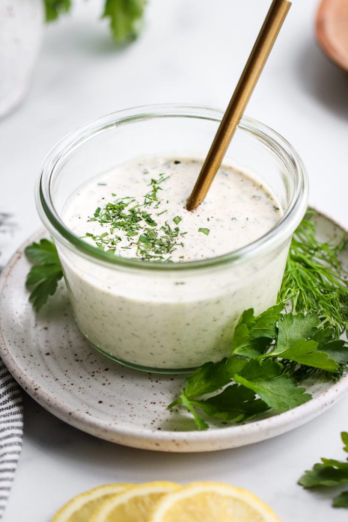 Paleo ranch dressing in a clear glass bowl sitting on a small speckled plate with fresh herbs scattered around.