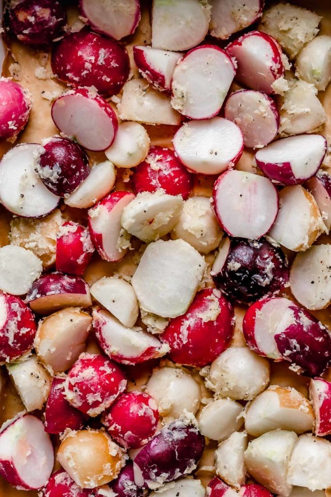 Overhead view raw radishes cut in half coated in oil and garlic on parchment-lined baking sheet.