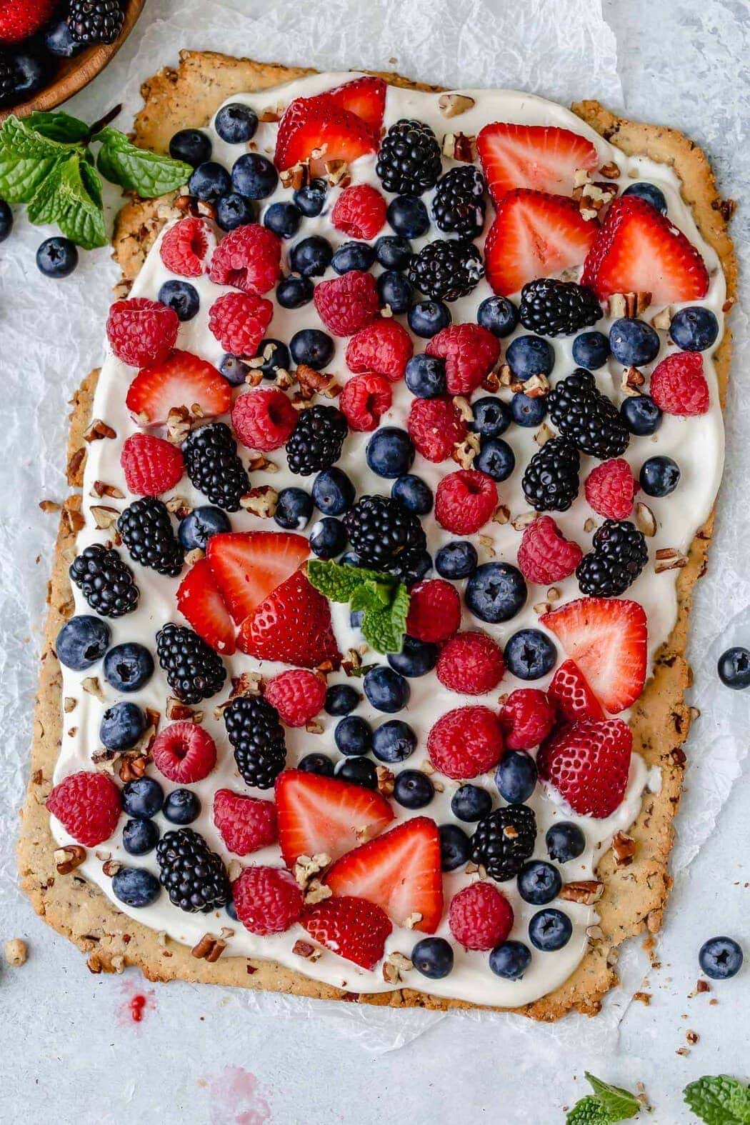 Overhead photo of Gluten-free Berry Fruit Pizza garnished with a spring of mint in the center. Pizza is topped with an assortment of berry, chopped pecans and a sprig of mint leaves.