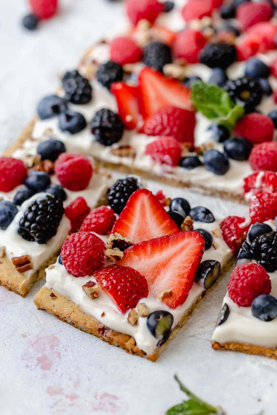 Photo of Gluten-free Berry Fruit Pizza garnished with a sprig of mint in the center. Pizza is topped with a Greek yogurt spread and an assortment of berries. This photo is showing the first row of pizza cut into squares. 