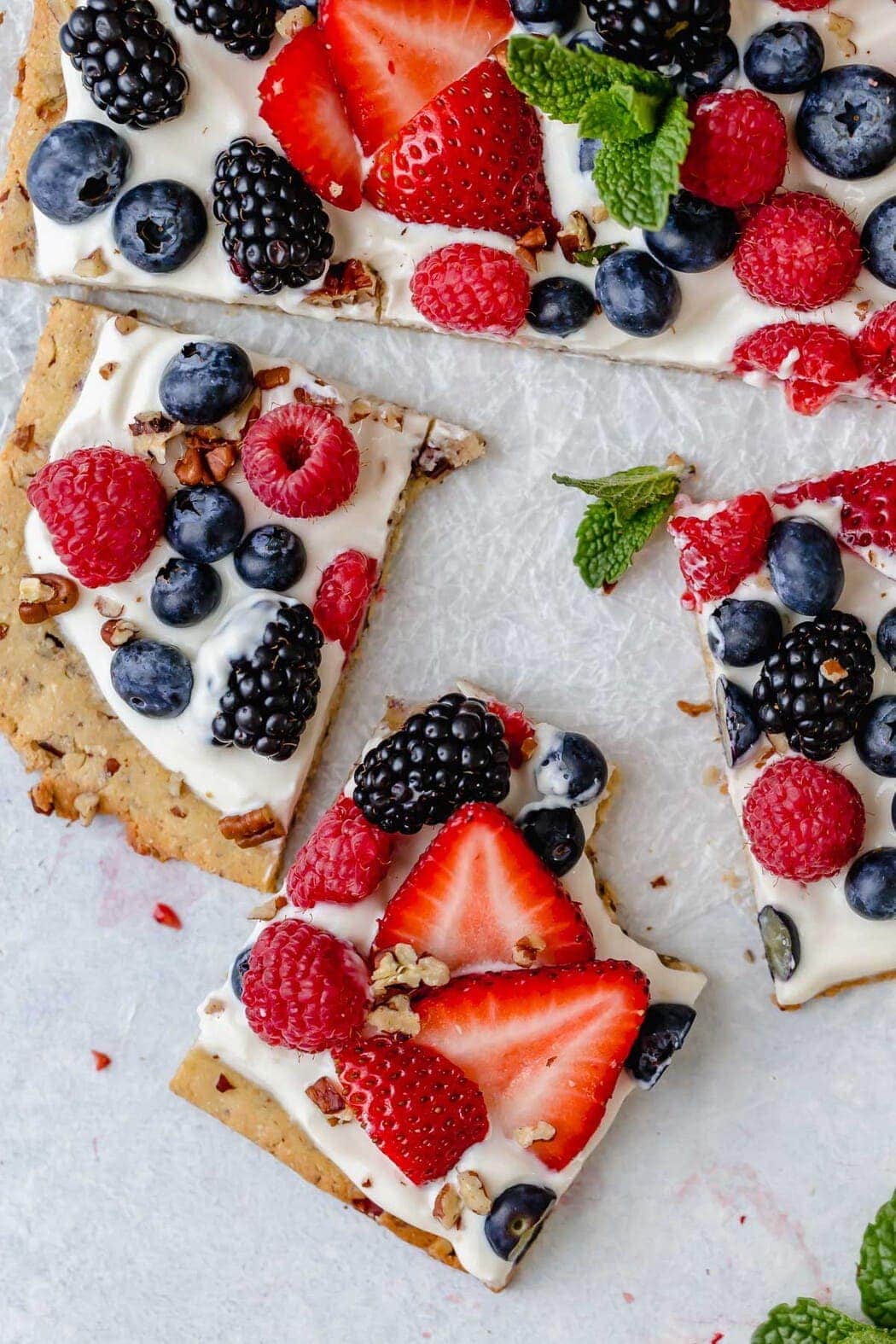 Overhead photo of Gluten-free Berry Fruit Pizza garnished with a sprig of mint in the center. Crust is topped with Greek yogurt and topped with an assortment of berries. Picture shows a bite taken out of one of the squares. 