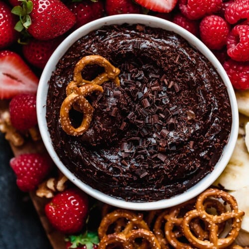 Overhead view of a white bowl filled with dark chocolate fruit dip dusted with chocolate shavings and surrounded by fresh fruit and pretzels.