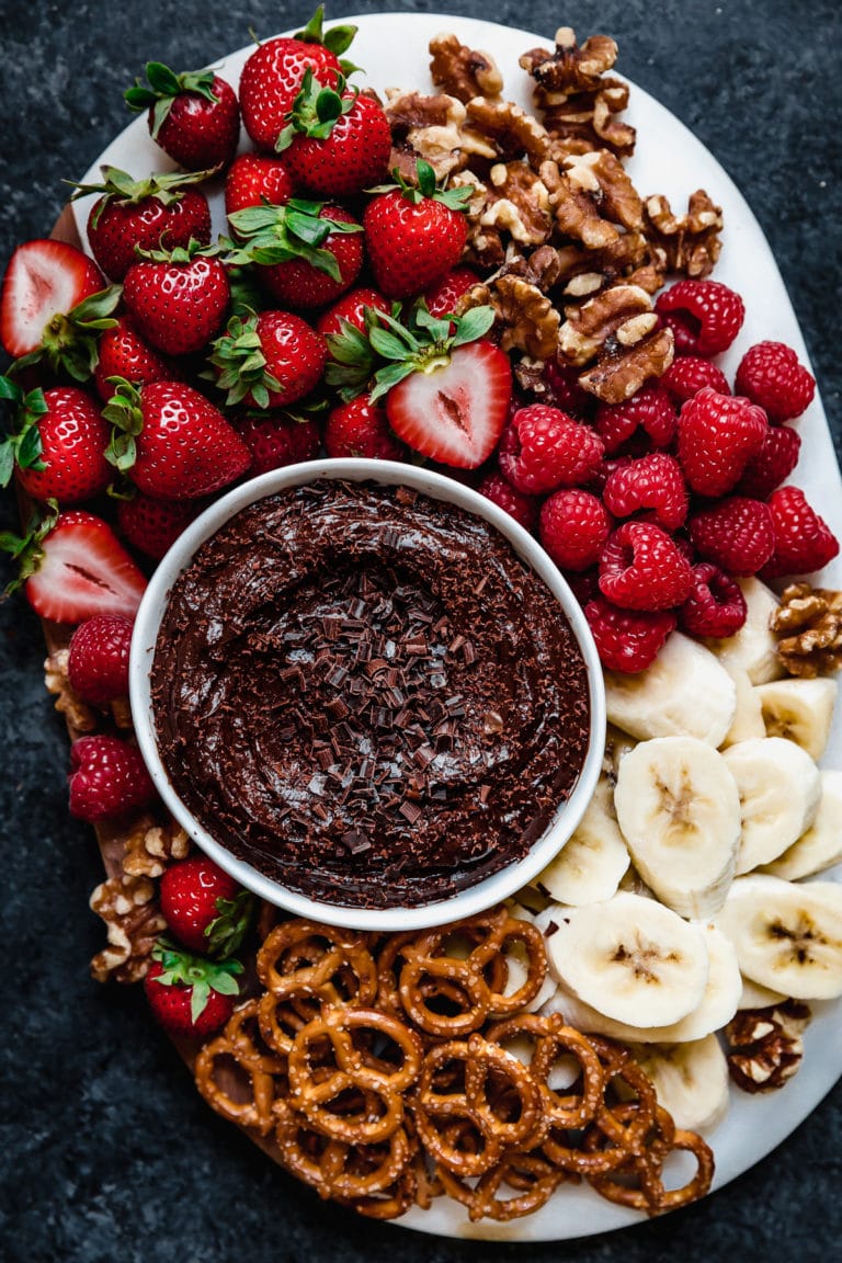 Overhead view white platter filled with fresh strawberries, banana slices, pretzels and pecans; surrounded a white bowl filled with chocolate avocado fruit dip