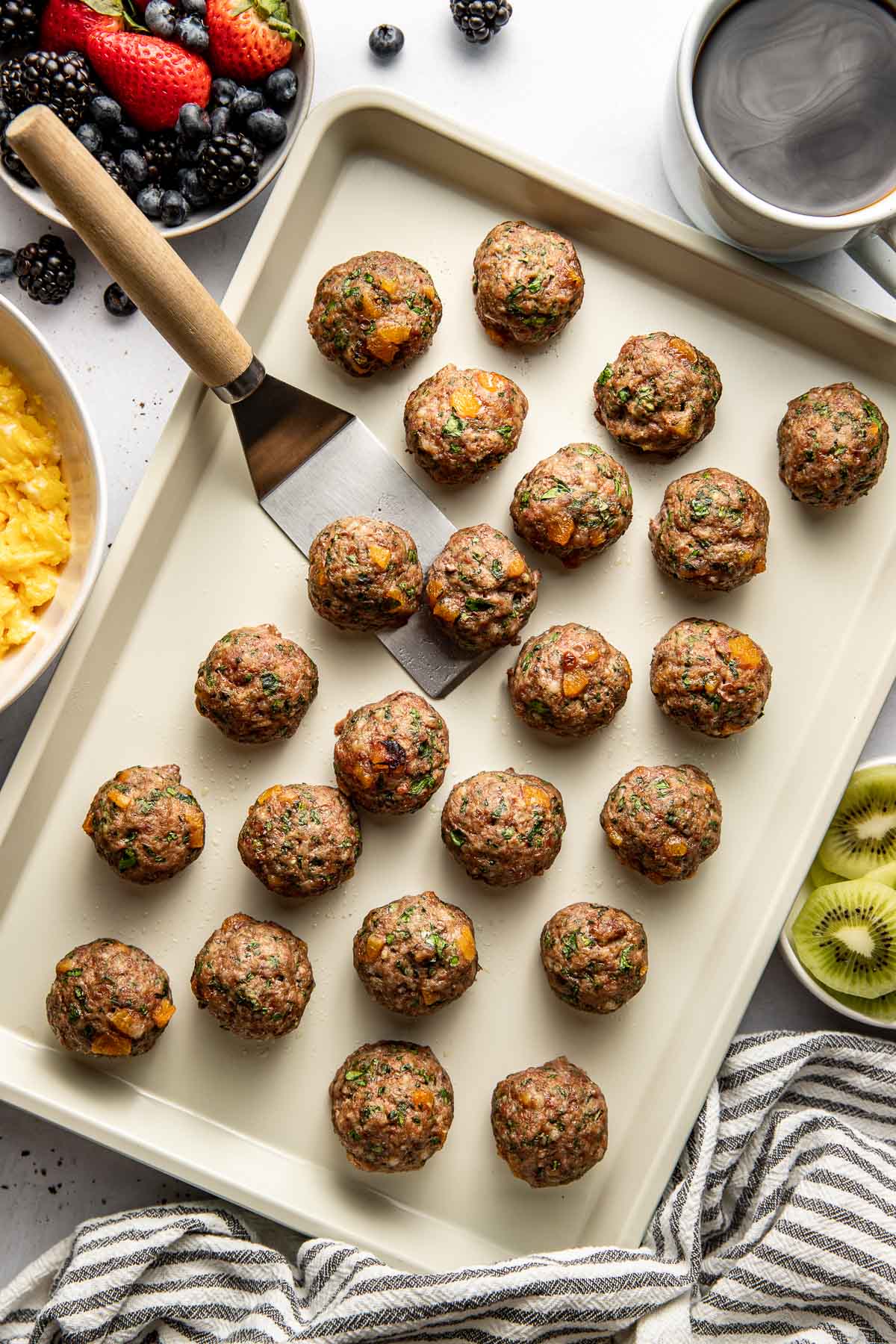 Overhead view of a sheet pan filled with Apricot Basil Breakfast Meatballs and a spatula scooping 2 of them. 