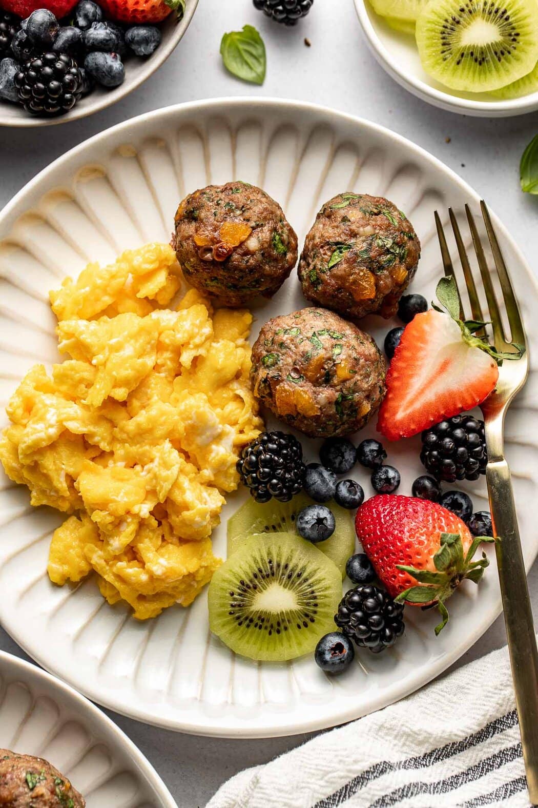 Close up view of a plate of Apricot Basil Breakfast Meatballs, scrambled eggs, and fresh colorful fruit. 