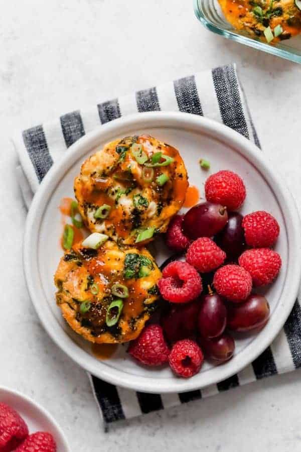 Two egg muffins served on white plate setting atop striped napkin, splashed with buffalo sauced and garnished with raspberries and grapes