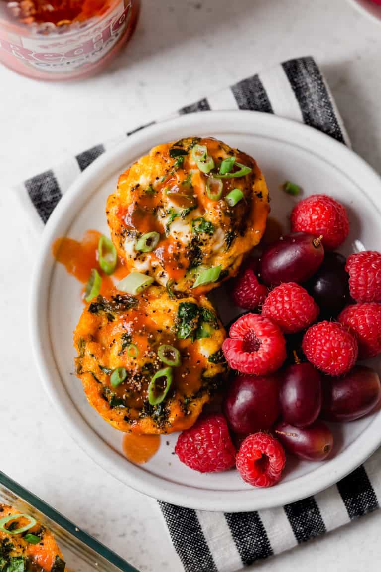Overhead view of a plate of buffalo chicken egg muffins topped with buffalo sauce and green onions next to fresh fruit. 