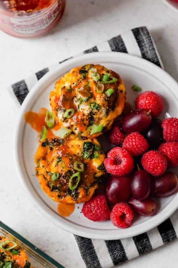Overhead shot of two Buffalo Chicken Egg Muffins on a white plate with a side of raspberries and grapes.