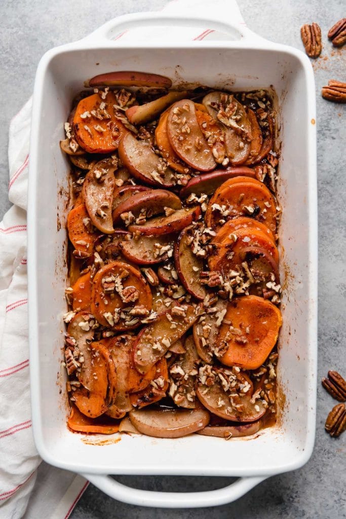 Overhead view white baking dish filled with cinnamon sweet potato apple bake topped with chopped pecans and maple syrup