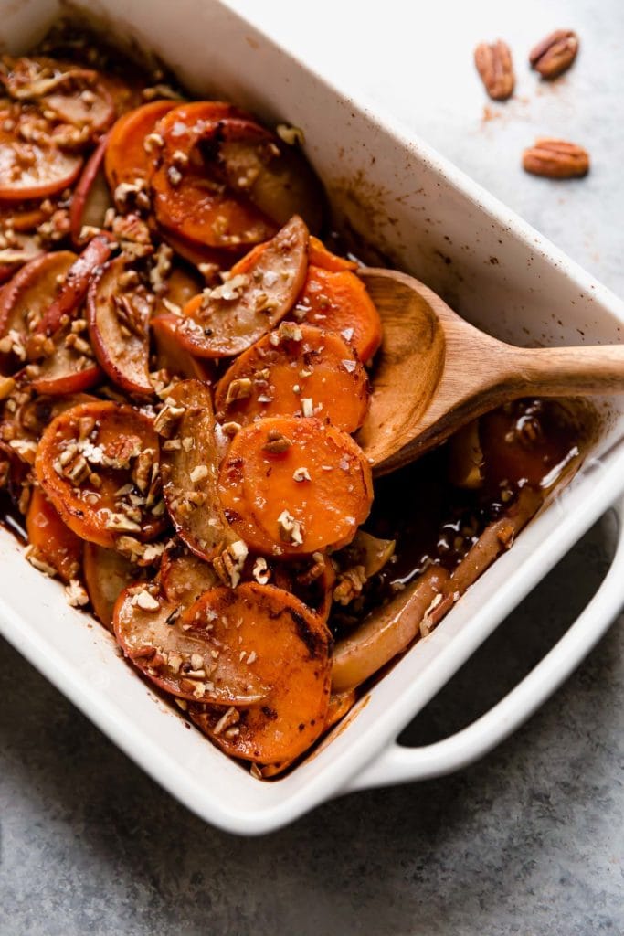 A wooden spoon scooping up serving of sweet potato apple bake with pecans from white baking dish