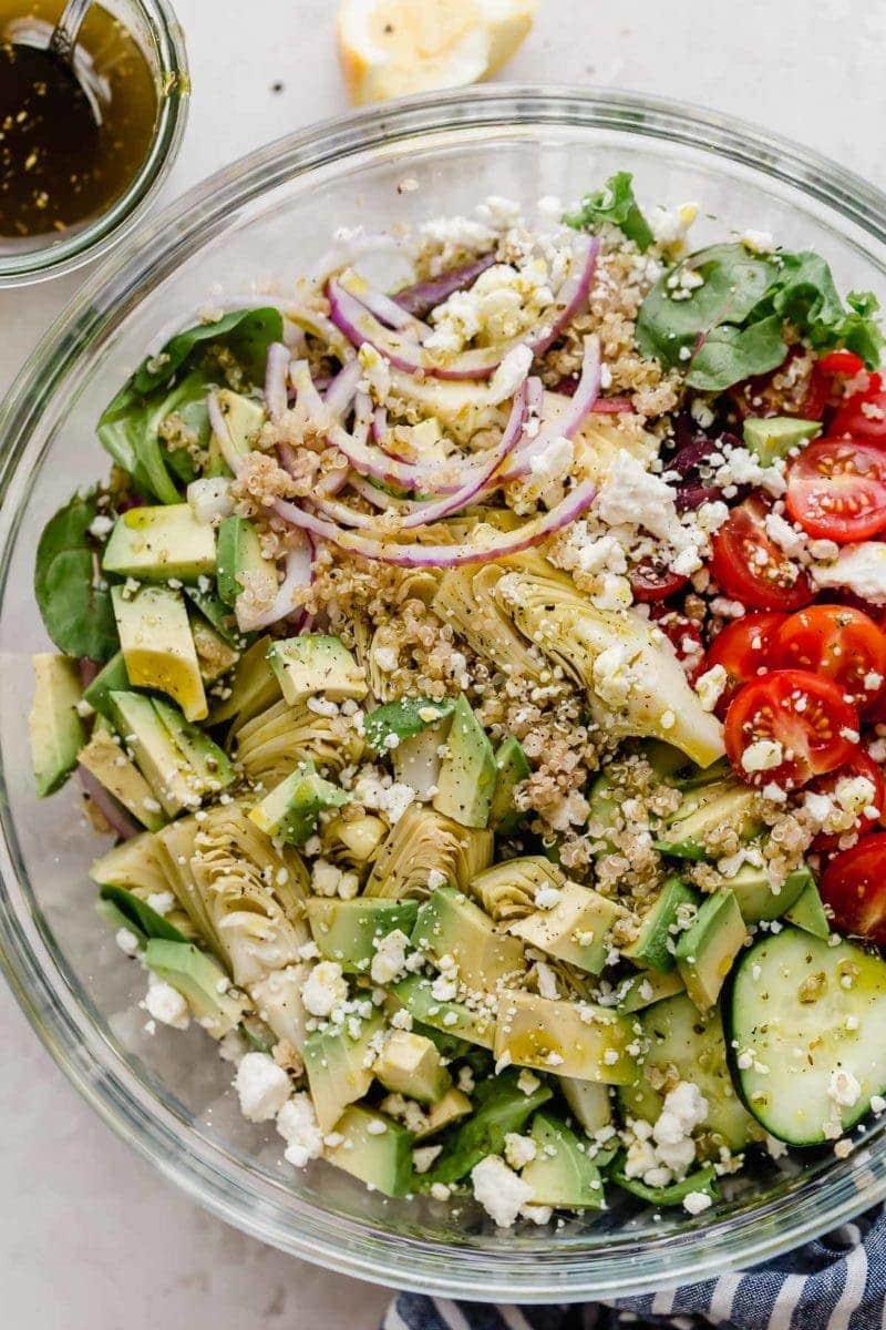 Close up view Greek Quinoa Salad with Avocado in a clear bowl before it has been mixed. Off to the side is the homemade vinaigrette in a small clear bowl.