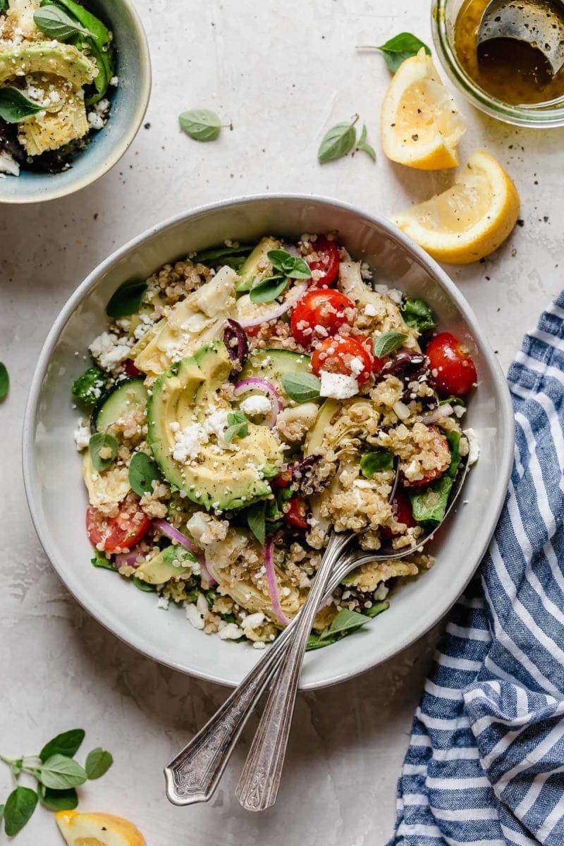 Photo of Greek Quinoa Salad with Avocado in a light gray bowl with serving spoons holding a scoop of the salad.
