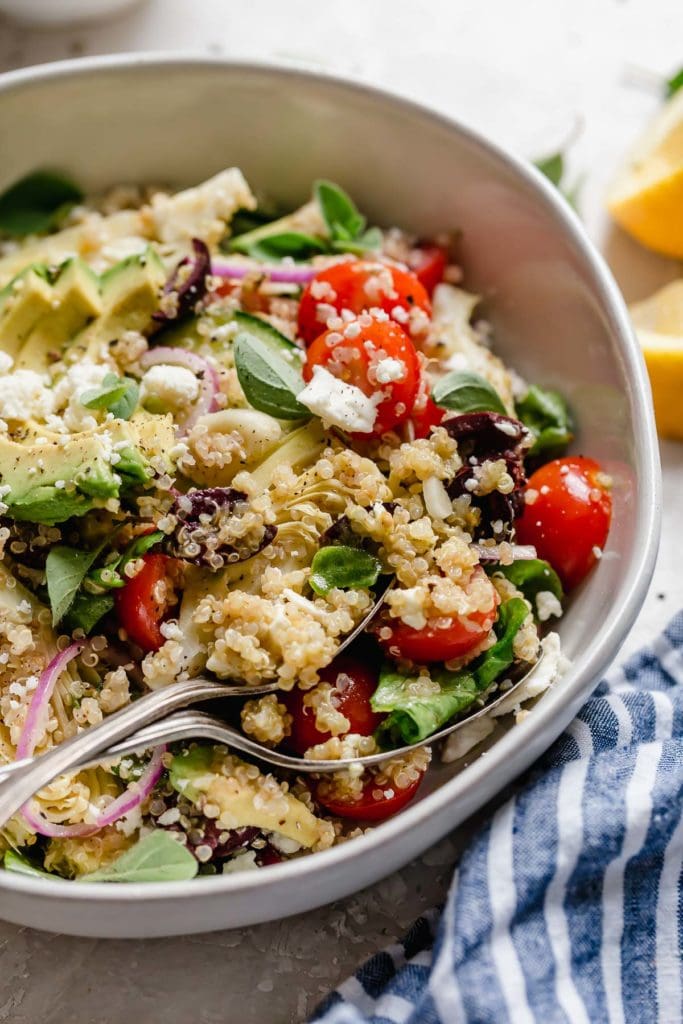 Greek Quinoa Salad with Avocado in a light gray bowl with serving spoons holding a scoop of the salad.