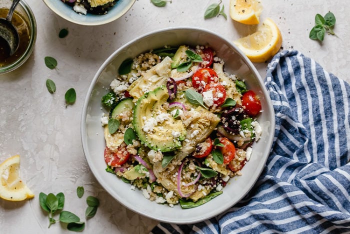 Greek quinoa salad with avocado slices and feta cheese served in white bowl topped with fresh herbs.