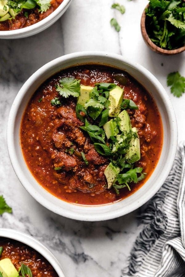 A white bowl filled with Slow Cooker Pumpkin Chili that's topped with diced avocado, fresh cilantro, and sliced green onions.